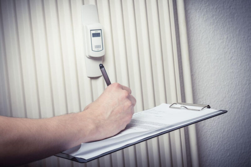 Rozliczanie kosztów centralnego ogrzewania w blokach ilustruje ręka pisząca na formularzu obok podzielnika ciepła na kaloryferze.